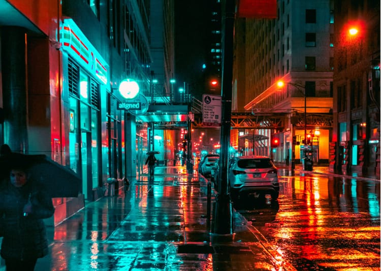 A wet, neon-soaked city street with reflections on the pavement and a lone pedestrian with an umbrella in the distance
