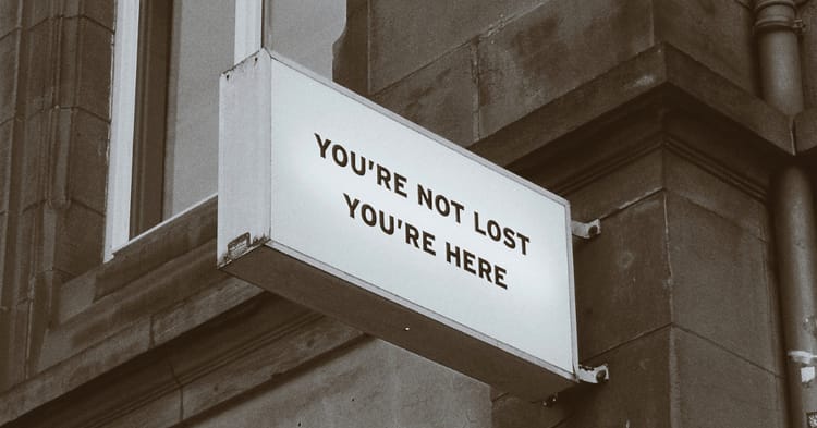 An oddly clear sign against a faded, grimy brick facade. The sign says YOU'RE NOT LOST YOU'RE HERE.