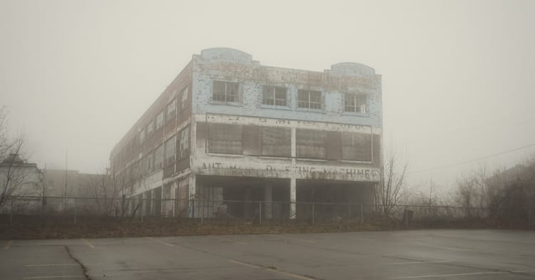 A rotting building with peeling paint stands in the fog with barren trees around it