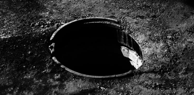 A broken circle of a manhole leading to dark shadow in a road scraped free of asphalt