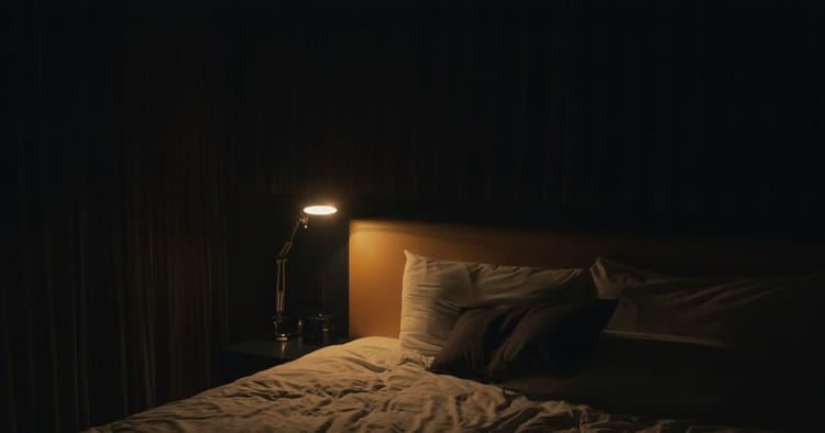 A single lamp casts a wan yellow light on a pillow and an empty bed in a pitch-black room