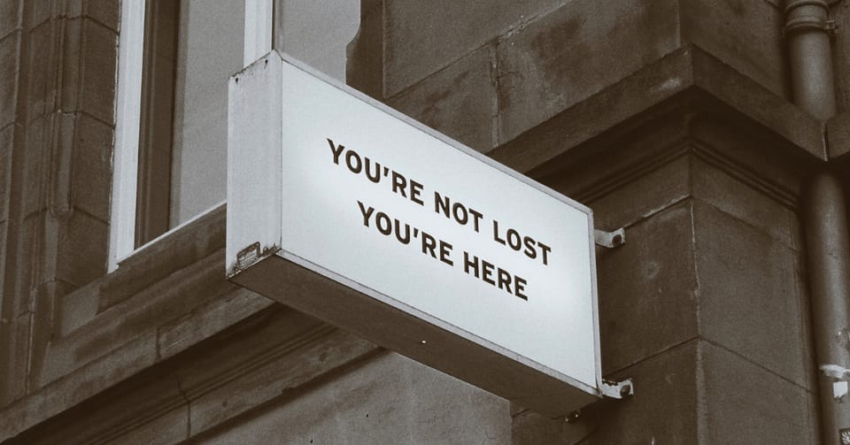 An oddly clear sign against a faded, grimy brick facade. The sign says YOU'RE NOT LOST YOU'RE HERE.