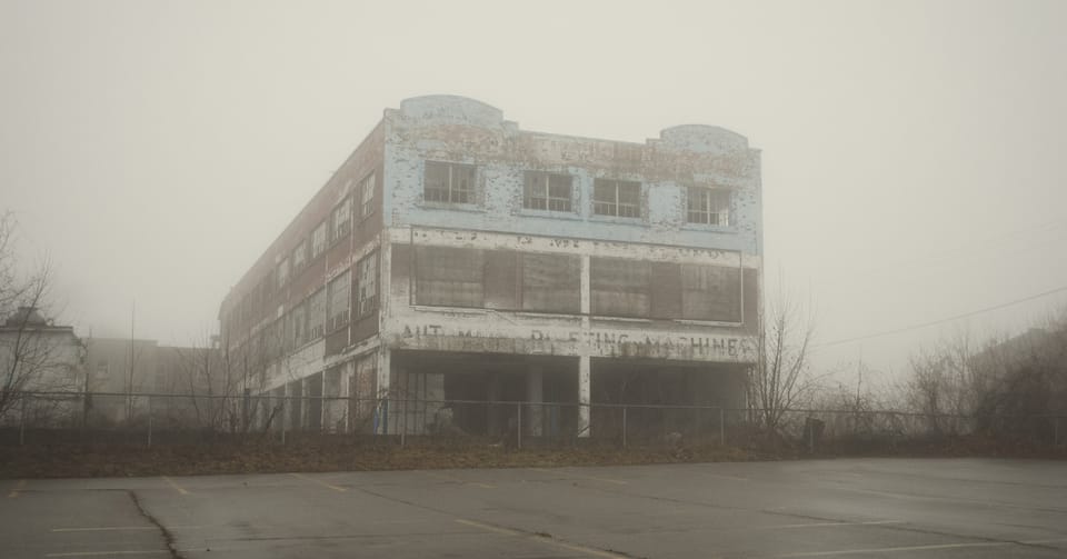 A rotting building with peeling paint stands in the fog with barren trees around it
