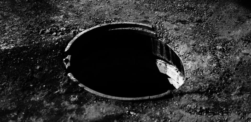 A broken circle of a manhole leading to dark shadow in a road scraped free of asphalt