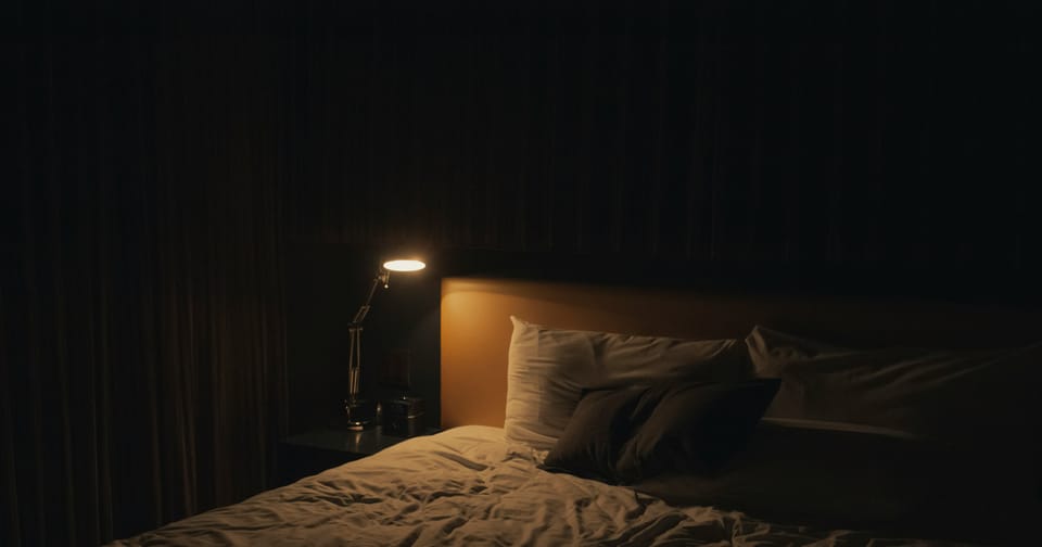 A single lamp casts a wan yellow light on a pillow and an empty bed in a pitch-black room